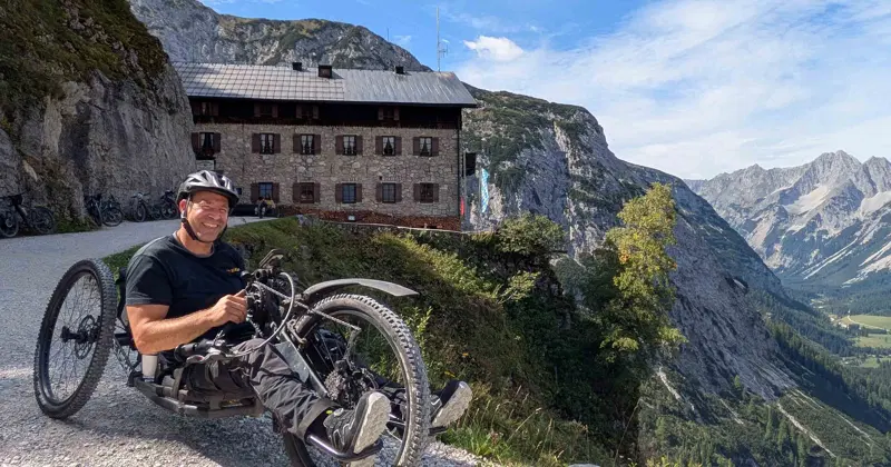 Een man op een ligfiets glimlacht op een bergpad van grind, met op de achtergrond een rustiek stenen gebouw en een indrukwekkend berglandschap.